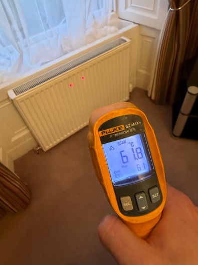 A hand holds an infrared thermometer reading 61.8°C, aimed at a white radiator