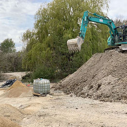 An excavator lifts dirt from a large mound at a construction site, surrounded by piles of sand and gravel under cloudy skies
