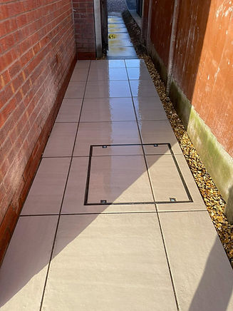 a narrow side alley paved with large beige or ivory porcelain outdoor tiles and a flush metal drain or manhole cover in the middle