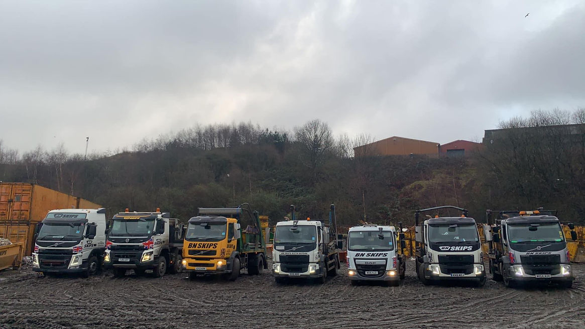 A row of skip trucks parked on a muddy lot, under a cloudy sky