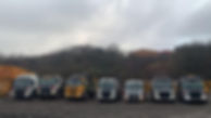 A row of skip trucks parked on a muddy lot, under a cloudy sky