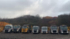 A row of skip trucks parked on a muddy lot, under a cloudy sky