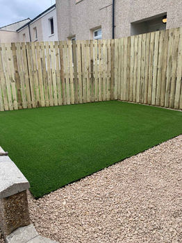 A small outdoor space featuring a green artificial grass area bordered by wooden fencing and stone gravel on one side
