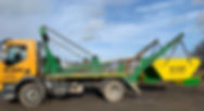 A yellow and green truck with a skip attached is parked on a dirt surface