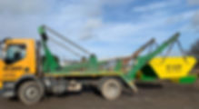 A yellow and green truck with a skip attached is parked on a dirt surface
