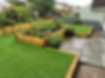 A landscaped garden featuring raised wooden planters with vibrant plants, lush artificial grass, and stone pathways