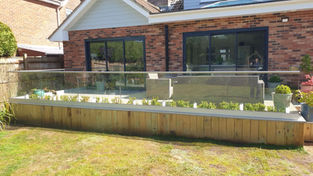 A wooden raised garden bed with young plants behind a glass balustrade, in front of a brick house
