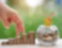 Fingers are placing a coin on a stack of ascending coins next to a glass jar