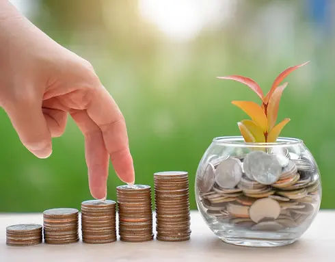 Fingers are placing a coin on a stack of ascending coins next to a glass jar 