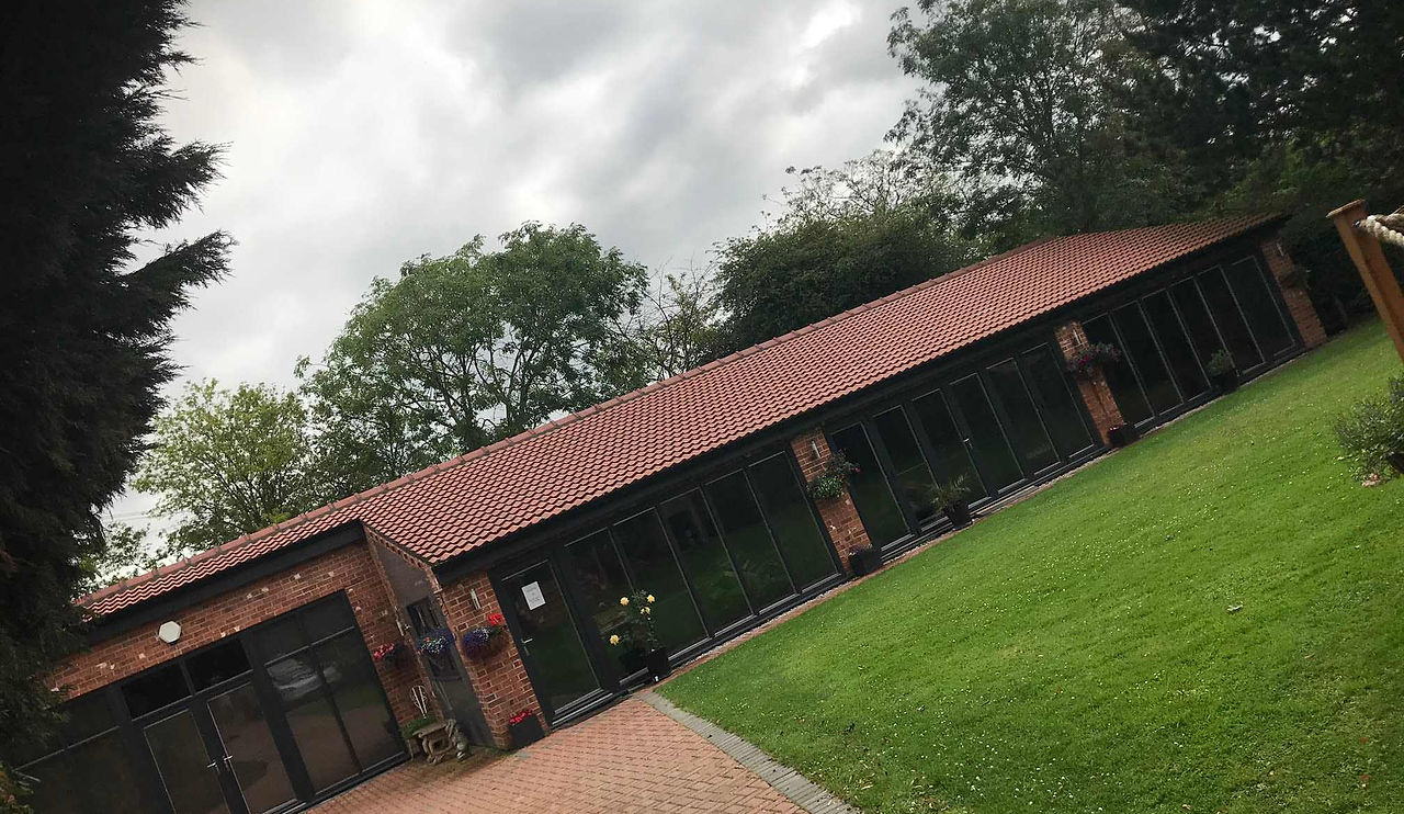 A modern single-story building with glass walls, a red tiled roof, surrounded by greenery and a paved pathway. Overcast sky above