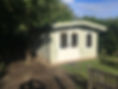 A small, light green garden shed with a white door and two windows stands on a brick base