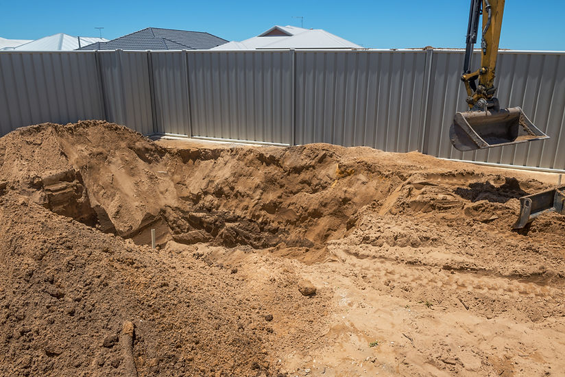 A large excavation pit in a sandy backyard and a yellow excavator arm on the left