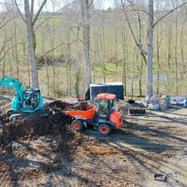 An aerial view of a construction site featuring an excavator and a dumper truck, surrounded by trees and construction materials