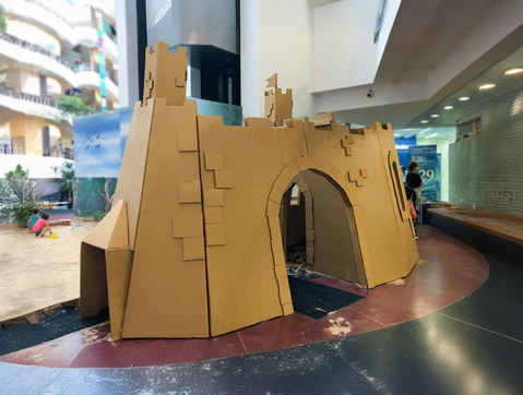 Large indoor walk-in castle structure made entirely of brown cardboard, featuring a central archway, battlements, and a sand play area on the left.