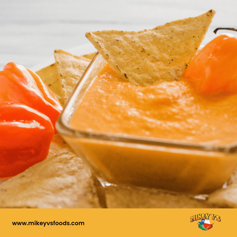 Glass bowl of orange dip with tortilla chips and a habanero pepper. Background shows light wood. Mikey V's Foods logo and site visible.
