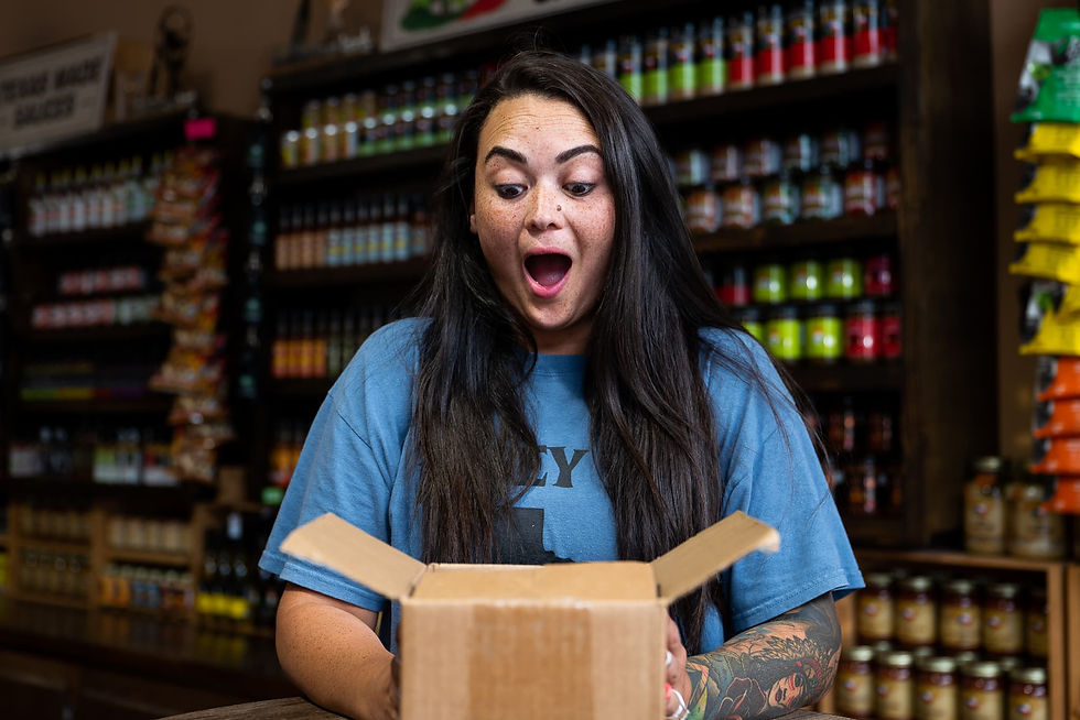 Woman in blue shirt, wide-eyed and surprised, looks into open box in a colorful shop with shelves of jars and snacks in the background.