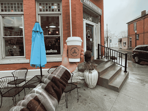 Hand holds takeaway coffee outside Espresso Air café on a rainy street in westerville, ohio; blue umbrella and red brick storefront.