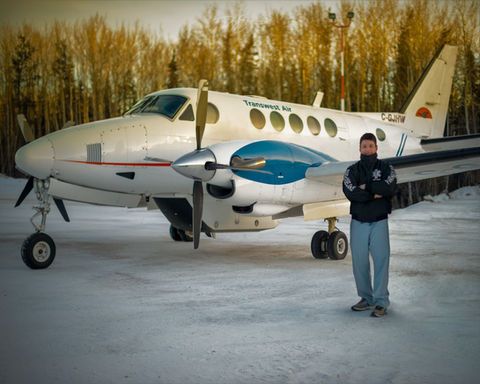 Dre Erwin, in scrubs, stands at the Pinehouse Lake air strip, prepared to provide compassionate healthcare to a client about to board a plane.