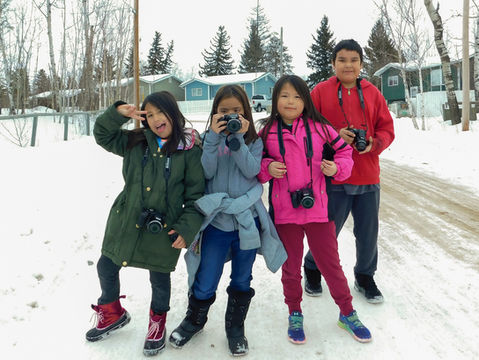 Four indigenous children standing in the snow holding up DSLR cameras and smiling for the camera.