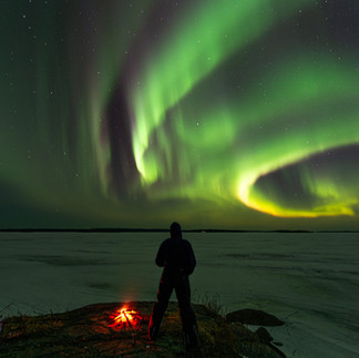 A lone spectator stands on a dark rocky shoreline next to a small, glowing campfire, looking out over a wide, frozen Saskatchewan lake beneath sweeping ribbons of green and purple northern lights.