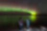 Two youths standing in awe, gazing at the Northern Lights reflected on Pinehouse Lake, experiencing the Aurora Borealis for the first time at night