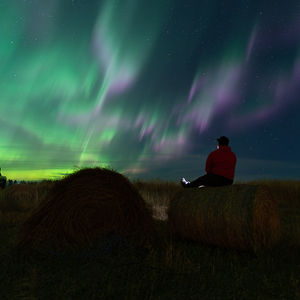 A man sits on a grain bale in a farmer's field near Regina, Saskatchewan, gazing at the mesmerizing Northern Lights in the autumn night sky. The auroras create vivid, swirling patterns of green and purple, providing a breathtaking spectacle