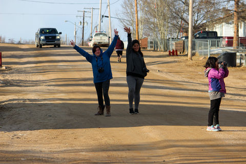 Two women waving cheerfully on a dirt road in a northern Saskatchewan community with a truck in the background.