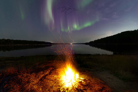 A bright campfire burning on a lake beach at night under a purple and green aurora borealis sky in Canada.