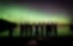 A silhouette of a group of youth standing with joined hands and raised arms along the shore of Pinehouse Lake, Saskatchewan. Above them, a vibrant green Aurora Borealis stretches across the starry night sky and reflects in the calm water. The scene features photography tripods, representing the Pinehouse Photography Club and its therapeutic photography mission to promote mental wellness and community connection.