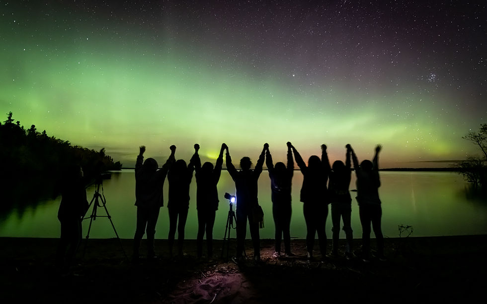 A silhouette of a group of youth standing with joined hands and raised arms along the shore of Pinehouse Lake, Saskatchewan. Above them, a vibrant green Aurora Borealis stretches across the starry night sky and reflects in the calm water. The scene features photography tripods, representing the Pinehouse Photography Club and its therapeutic photography mission to promote mental wellness and community connection.