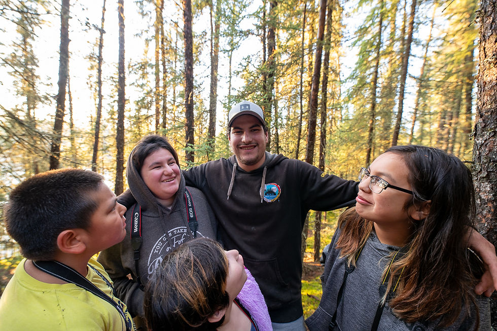 Group of five people, smiling in a forest with sunlight filtering through trees. Casual attire, relaxed mood, nature background.