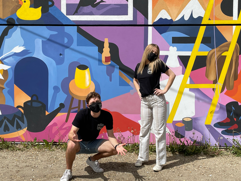 Masked man crouches, woman stands, posing before vibrant abstract mural.
