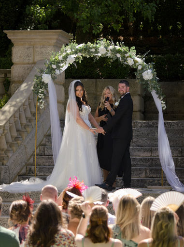 A bride and groom holding hands at the altar under a white floral arch during an outdoor wedding ceremony, captured by Perth wedding photographer Castle Media at Caversham House.