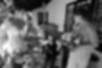 Candid black and white shot of a young page boy sitting in a wicker chair, surrounded by bridesmaids on a sunlit veranda, by a Perth wedding photographer.