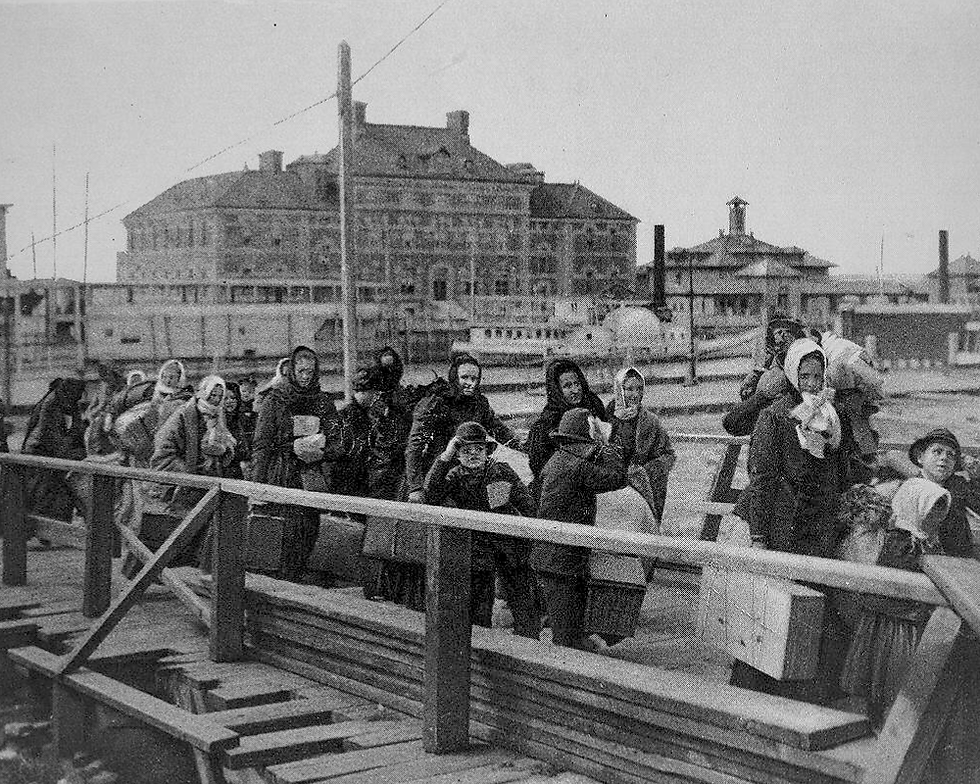 A black and white photograph from 1902 showing a crowd of European immigrants carrying luggage and walking down a wooden gangplank at Ellis Island, with historical brick terminal buildings in the background."