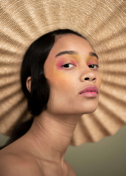A photograph of a female model, wearing a large straw hat, with pink lipstick, photographed against a pale moss green background