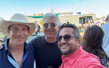 Three men smiling outdoors at a sunny gathering.