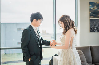 Katy and Obe holding hands by a window in formal wedding attire.