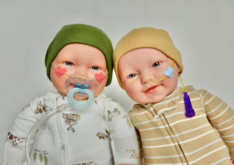 Two baby dolls sitting next to each other, one wearing green and the other yellow striped onesie. One doll has an oxygen cannula and the other a feeding tube