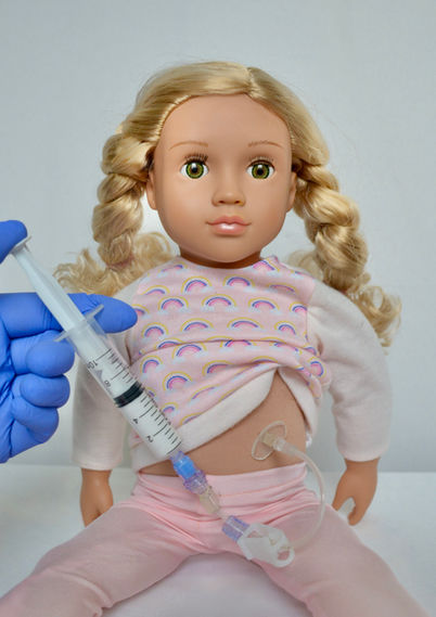 A blonde girl doll with a G-tube receiving a feed through a syringe