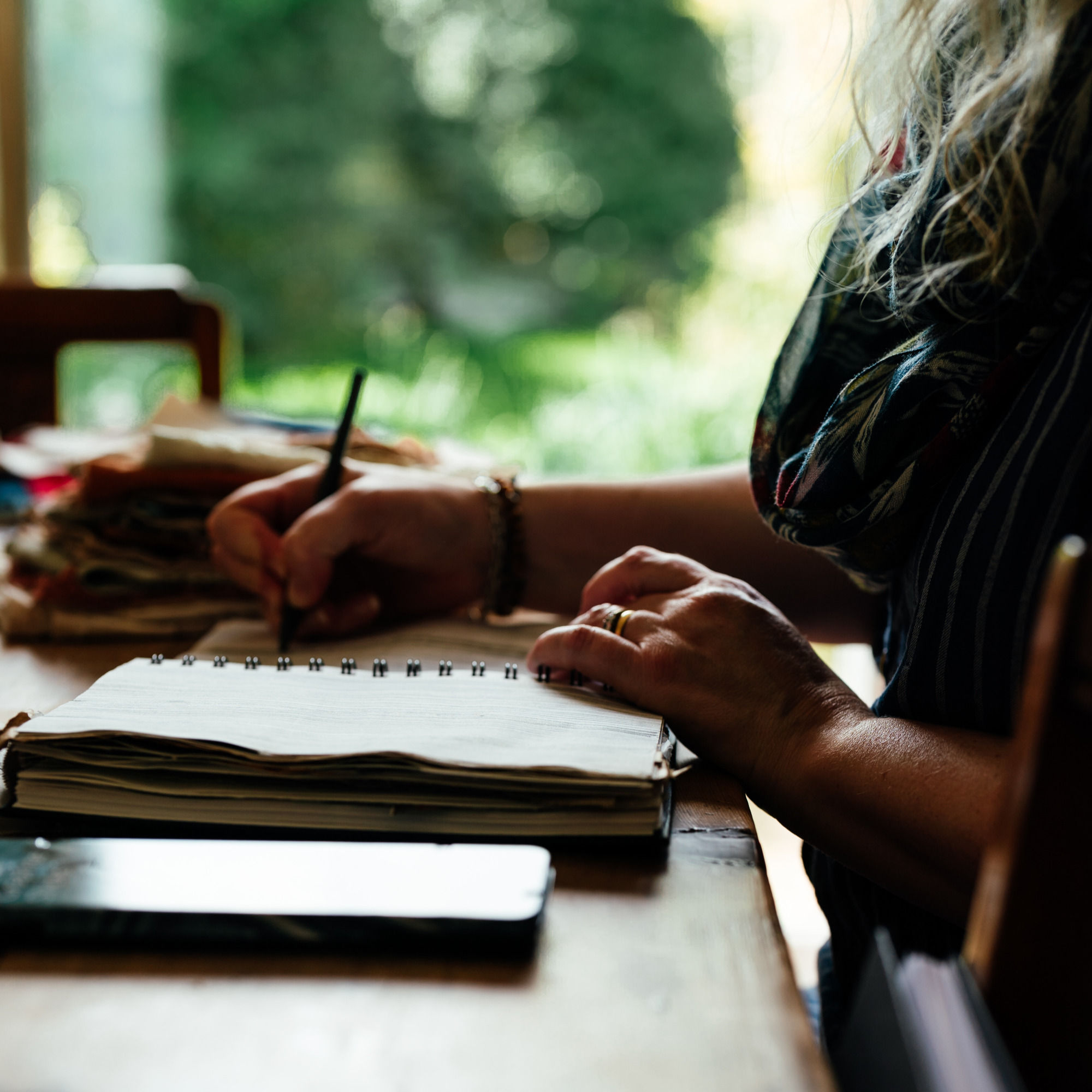white woman writing in a book sitting at a table in front of a window