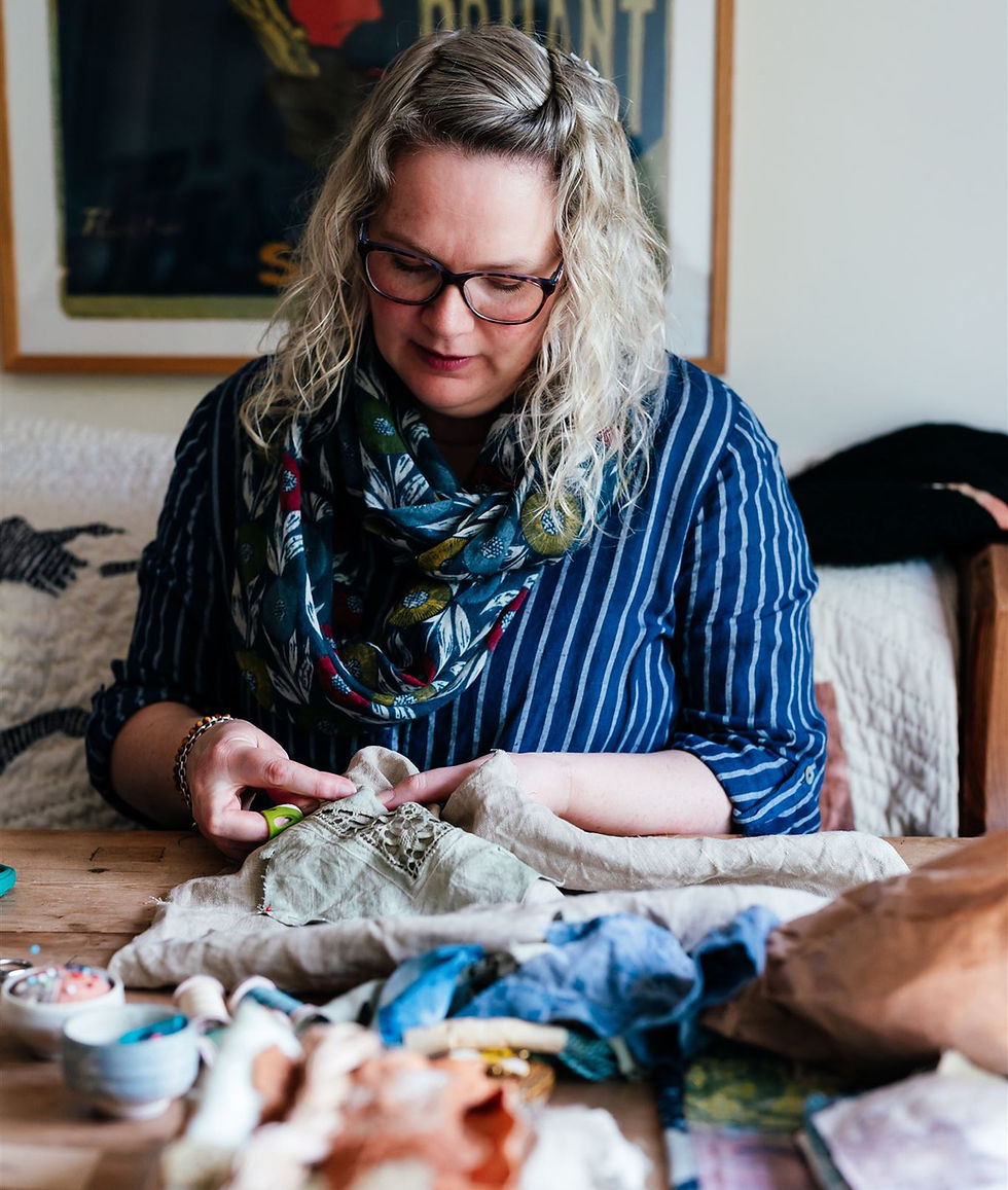Portrait by Holly Bobbins Photographer.
White woman in a blue dress with white stripes, sitting at a table, sewing by hand.