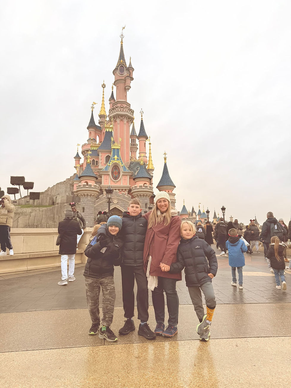 Four people pose happily in front of a pink castle at Disneyland. They're dressed in winter clothes. The atmosphere is cheerful and busy.