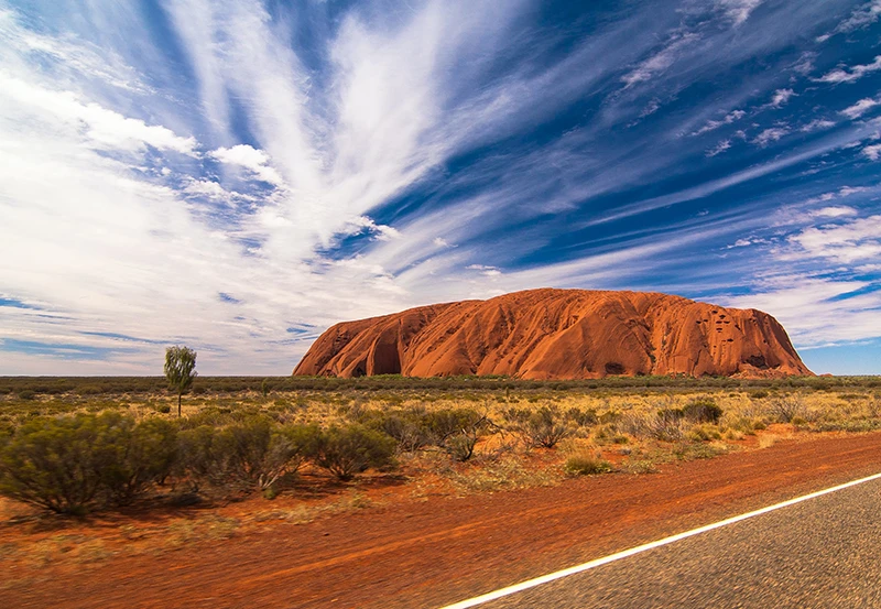 a desert landscape with a large rock formation in the distance with Uluru in the background