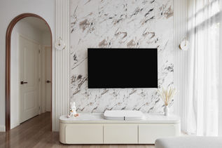 Close-up view of the luxurious TV wall feature in a modern luxe condo designed by Comfort Home Interior. The wall is clad in stunning marble tiles with intricate veining that adds depth and texture, flanked by vertical fluted panels that create a refined and sophisticated look. A sleek, minimalist TV console sits below, continuing the marble pattern for a cohesive design. The arched doorway and soft, ambient lighting fixtures add a touch of warmth and elegance to the space, making this wall feature a true centerpiece of the living area, blending classic luxury with modern design sensibilities.