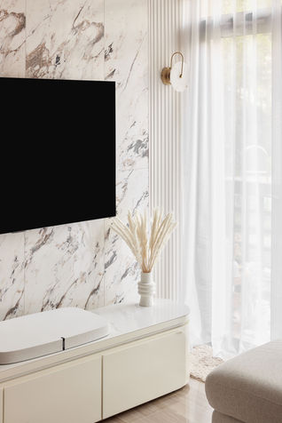 The focal point is a stunning marble wall feature, characterized by intricate veining and a polished finish. The marble panels are seamlessly integrated with vertically grooved details on either side, adding a touch of elegance and texture to the space. To the right, soft, sheer curtains filter natural light into the room, creating a serene and airy atmosphere. The decor is minimalist, with a sleek, curved TV console in a neutral tone positioned below a wall-mounted flat-screen TV. A simple vase with pampas grass adds a natural element to the refined setting.