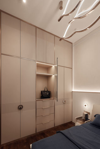  A modern luxe master bedroom designed by Comfort Home Interior, showcasing a custom-built wardrobe with integrated lighting. The wardrobe features a mix of closed storage and open display shelves, accented by gold-finished handles and trim. The bed, with a textured headboard and neutral linens, completes the sophisticated look of the room.