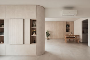 A minimalist 4-room resale flat in Bishan, Singapore, designed by Comfort Home Interior, showcasing a clean and serene living space. The design features custom-built cabinetry in neutral tones, seamlessly integrated with the architecture of the home. The absence of lighting enhances the natural, subdued ambiance, emphasizing the simplicity and elegance of the mid-century modern aesthetic. The curved entrance wall transitions smoothly into an open dining area, where a wooden dining table with matching benches invites warmth and functionality. The space is designed with an emphasis on natural light, uncluttered surfaces, and a harmonious blend of form and function, characteristic of minimalist interior design.