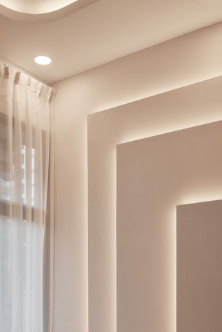 This image captures the feature wall in a modern luxe condo designed by Comfort Home Interior. The wall features an intricate stepped design with subtle textures, illuminated by soft recessed lighting that accentuates the layers, creating depth and a sense of elegance. The smooth transition between the wall and ceiling is complemented by sheer curtains that allow natural light to filter into the space, adding a warm, welcoming ambiance to the room. The wall seamlessly integrates with the overall aesthetic of the living area, contributing to the clean, sophisticated look of the space.