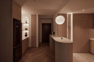 Japandi-style condo kitchen and entryway illuminated by warm ambient lighting, highlighting the natural wood finishes and minimalist design. The curved countertop and built-in shelving add to the serene and functional layout. The space is enhanced by soft lighting that creates a cozy, inviting atmosphere, perfect for evening relaxation.